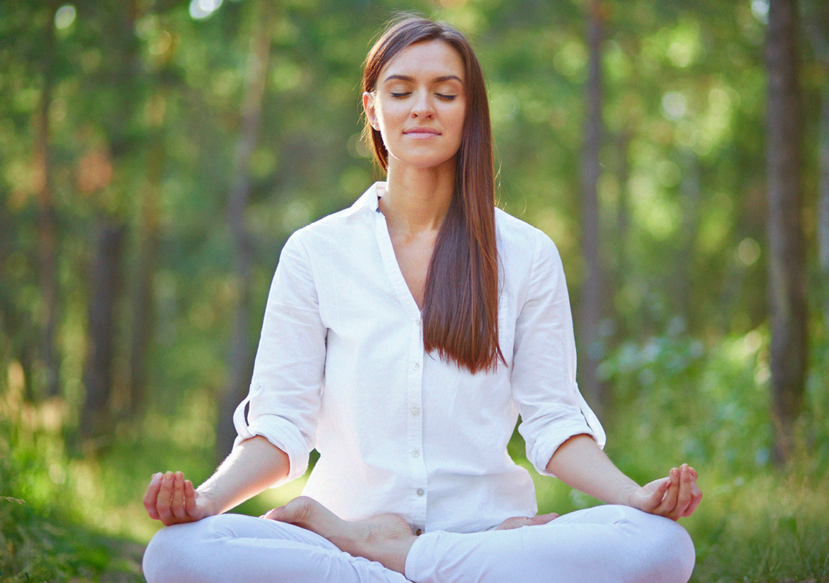 Mulher levemente sorrindo com roupas brancas leves sentada no gramado em posição namastê (de pernas cruzadas e com as palmas das mãos encostadas uma na outra) praticando meditação.