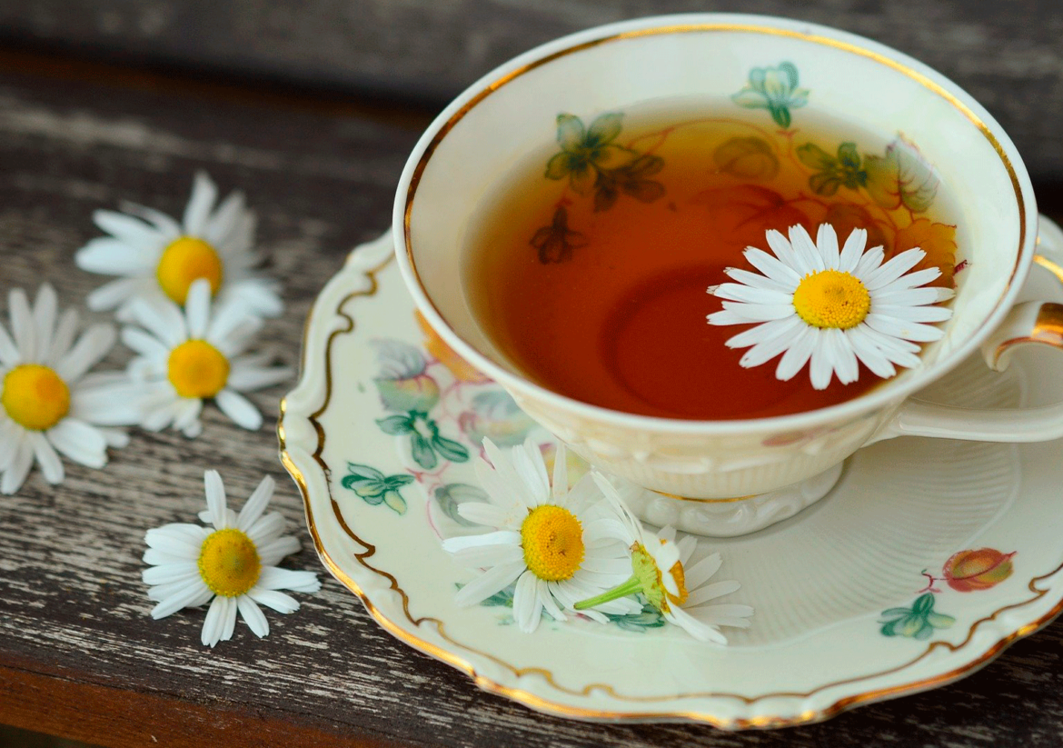 Xícara de porcelana com desenhos floridos, com chá e flores da camomila, em cima de uma mesa de madeira - Chá de camomila é um dos alimentos que pode ajudar na qualidade do sono