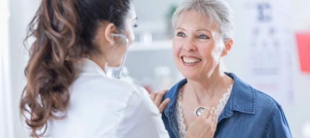 Mulher idosa (de frente) sorrindo para médica (de costas) que está medindo os batimentos do coração.